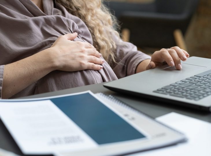 Close-up of pregnant businesswoman touching her belly while sitting at the table and typing on laptop during online work at office